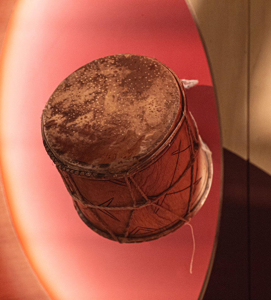 Tambor de madeira natural clara com grafismos marrons, amarrações em corda fina e couro na parte superior onde se produz o som. 