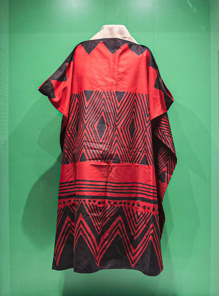 Veste longa, vermelha, com triângulos, listras e pontos pretos formando grafismos regulares por toda a extensão do tecido.