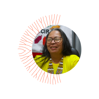 Mulher indígena, cabelos longos com mecha branca na frente, usa óculos, uma blusa amarela, um colar de contas coloridas e sorri.