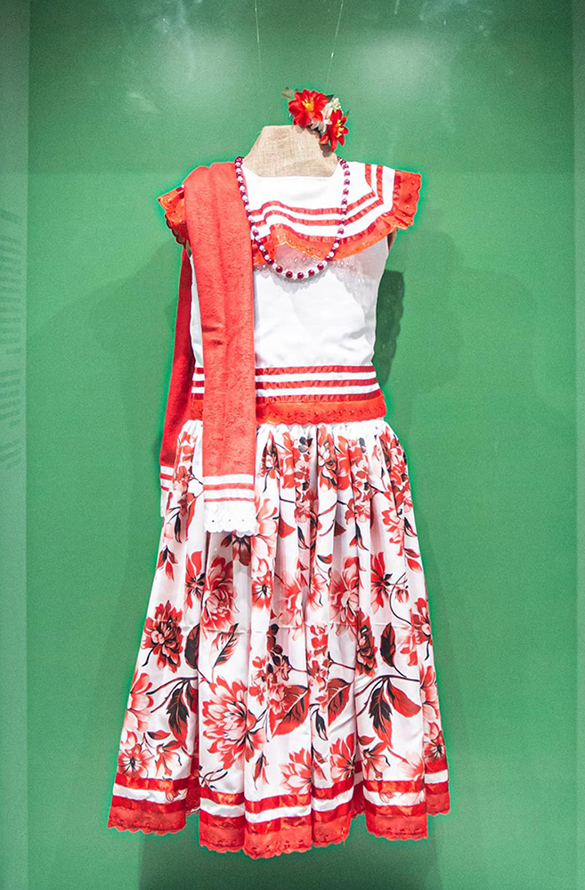 Vestido branco com estampa de flores vermelhas na saia e blusa lisa, enfeitado na barra, gola e cintura com fitas e entremeios vermelhos. 
